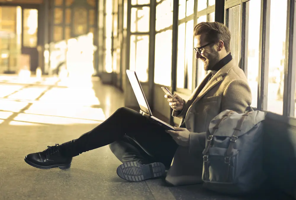 Un homme souriant travaillant à distance avec son ordinateur portable et son smartphone, assurant la gestion des centres de formation certifiés Qualiopi.