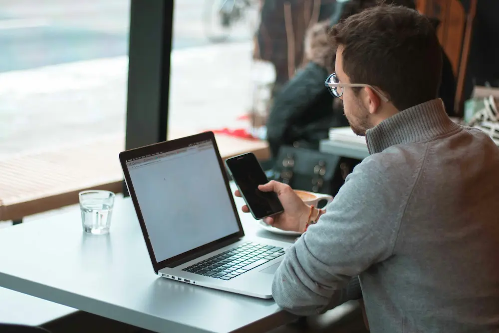 Un homme utilisant son smartphone tout en travaillant sur un ordinateur portable dans un café.